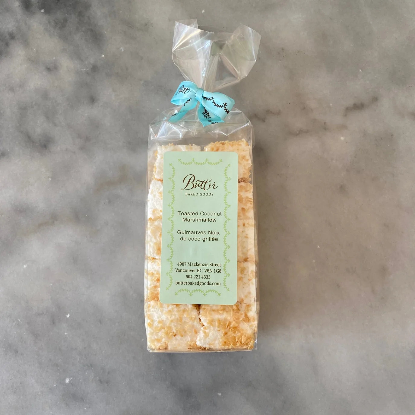 A packaged loaf of toasted coconut marshmallows on a gray marble surface. The cookies are visible through a clear plastic bag tied with a light blue ribbon, with a pale green label on the front reading 'Bour' and 'Toasted Coconut Marshmallow'.