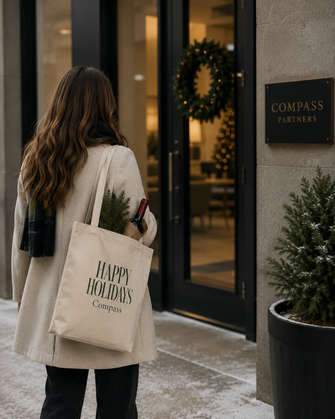 A woman in a beige coat stands in front of a building entrance carrying a cream canvas tote bag printed with 'HAPPY HOLIDAYS Compass' in dark green text. The scene includes a holiday wreath on the glass doors, a small potted evergreen, and a wall sign reading 'COMPASS PARTNERS'.