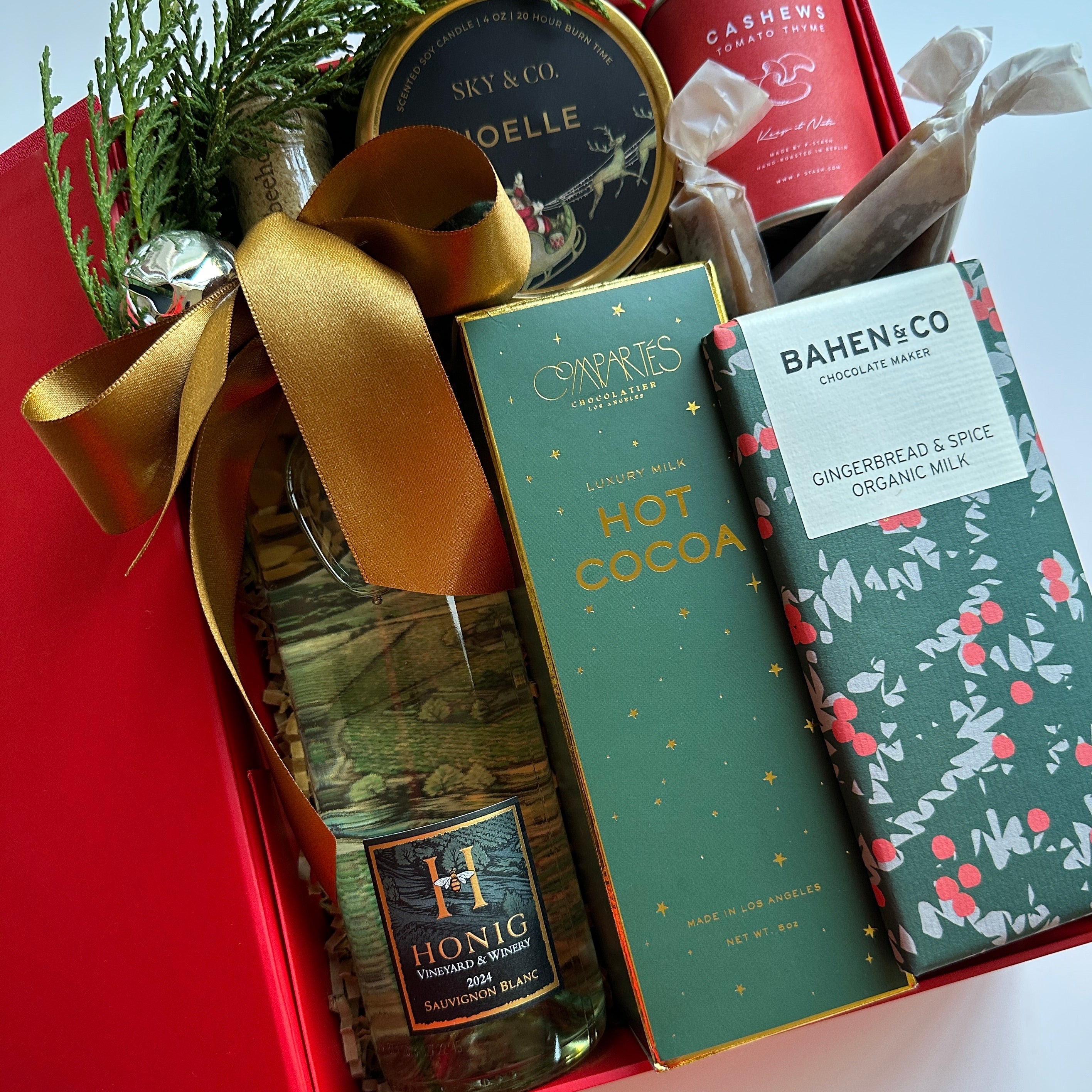 Gift box with chocolate bars, a bottle of alcohol, and a canister, decorated with a gold ribbon on a light blue background.