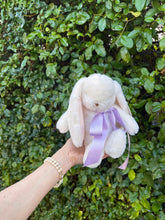 Hand holding a plush bunny with a purple bow against a green leafy background