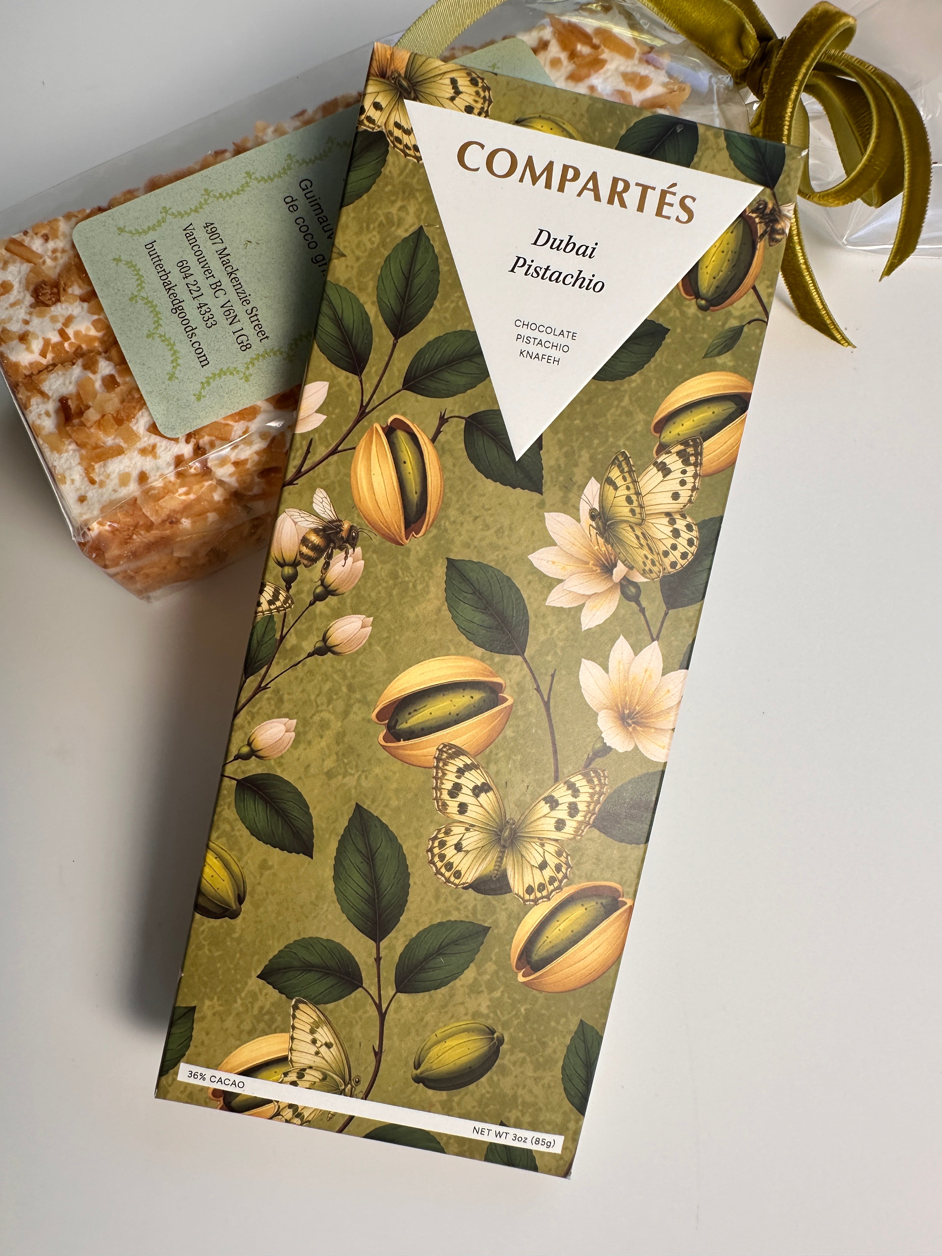 A packaged chocolate bar labeled COMPARTÉS Dubai Pistachio, shown with its illustrated box in front of a bag of toasted marshmallows.