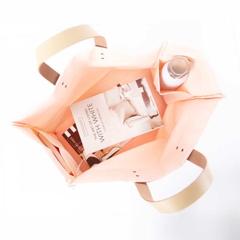 A light blush-pink fabric tote bag with beige handles, shown from a top-down angle on a white background, filled with various items including a book and a bottle.