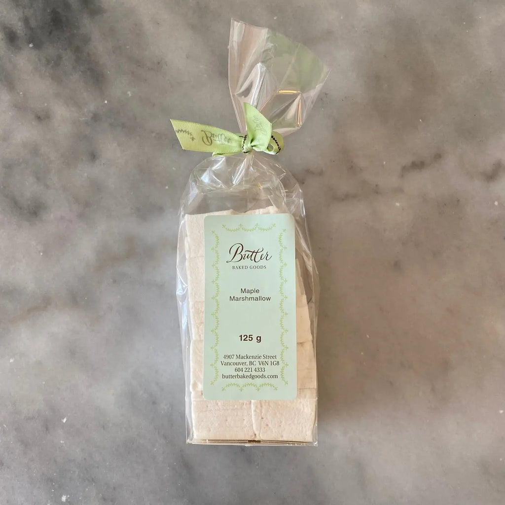 A packaged loaf of Butter Bakery's Maple Marshmallows, wrapped in clear cellophane with a light green ribbon and a pale green label, placed on a gray marble surface.