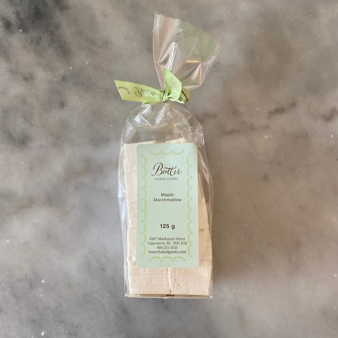 A packaged loaf of Butter Bakery's Maple Marshmallows, wrapped in clear cellophane with a light green ribbon and a pale green label, placed on a gray marble surface.