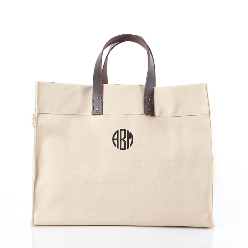 A beige canvas tote bag with dark brown leather handles and a black circular monogram logo on the front, set against a plain white background.