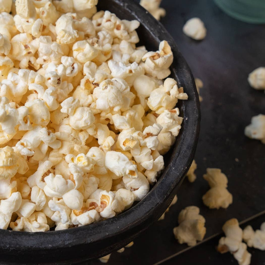 A close-up of fluffy white popcorn filling a dark bowl, with a few popcorn pieces scattered on a dark surface.