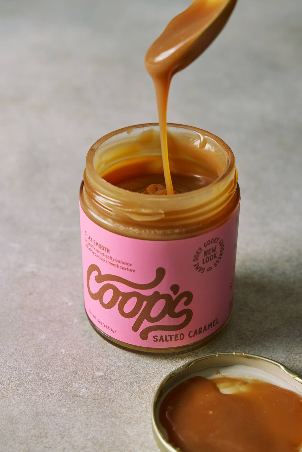 Jar of Coop's salted caramel with a spoon pouring caramel into it on a neutral background