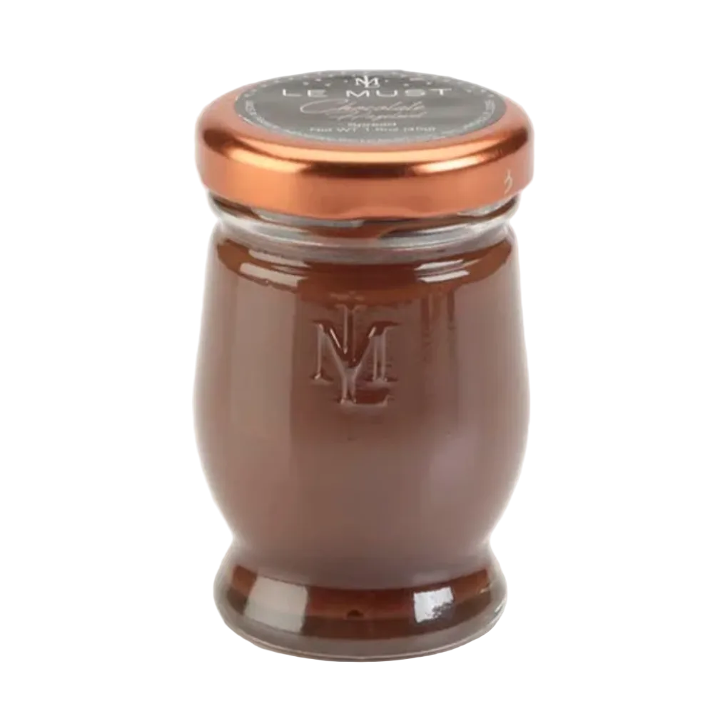 A glass jar  with a metallic copper-colored lid and a dark gray label reading 'Le Must', filled with hazelnut spread and featuring an embossed 'LM' logo on the front, set against a plain white background.