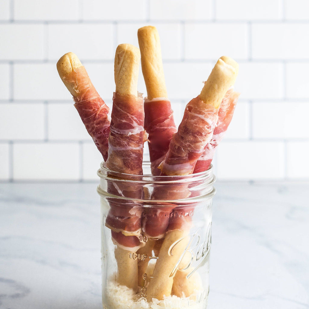 Glass jar filled with bread sticks wrapped in prosciutto on a white tiled background