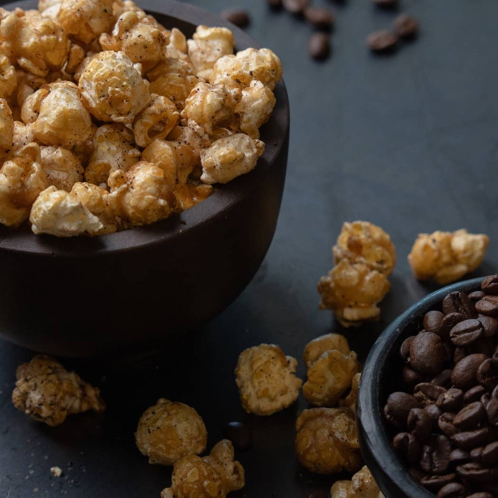 Caramel popcorn in a dark bowl with scattered caramel popcorn pieces and a small bowl of coffee beans on a dark surface.