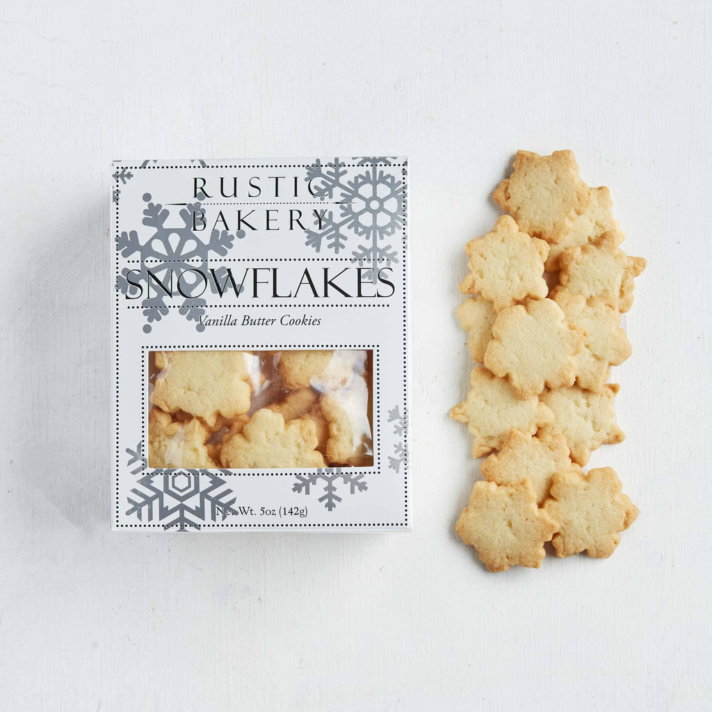 A box of Rustic Bakery Snowflakes vanilla butter cookies sits on a white surface next to several baked snowflake-shaped cookies.