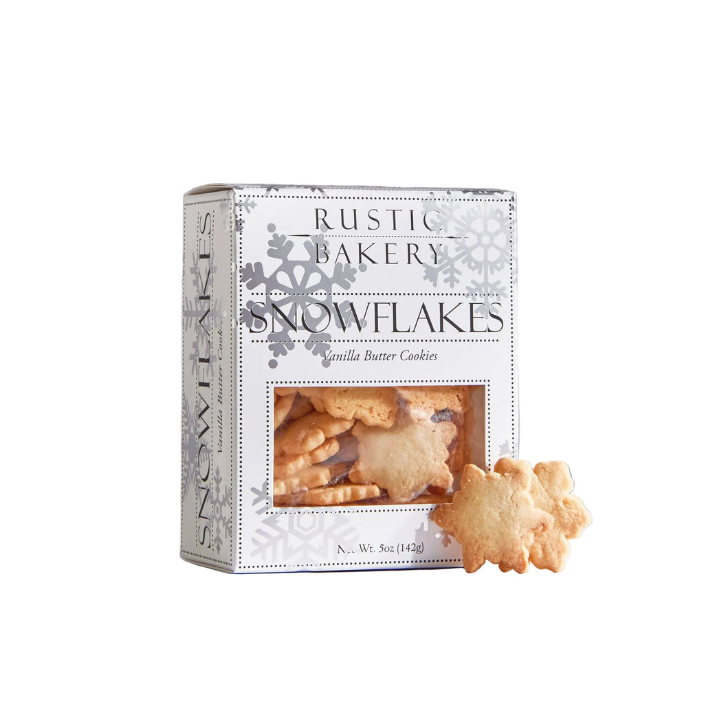 A box of Rustic Bakery Snowflakes vanilla butter cookies stands on a white surface against a light gray background, with two golden-brown snowflake-shaped cookies placed in front of the package.